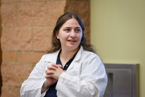 A female instructor wearing a white lab coat with hands clasped in front of them looks to the right.